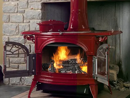 Red cast iron wood stove with glass doors, burning wood, and a warming oven on top.